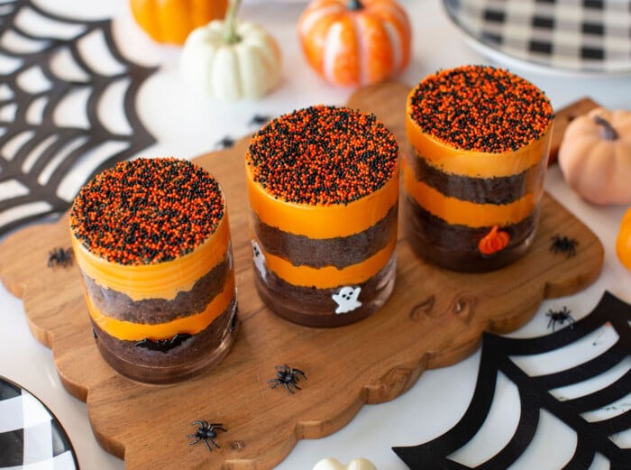 Three glass jars filled with layered orange and brown Halloween desserts, topped with orange and black sprinkles, sit on a wooden board. Small plastic spiders and festive pumpkins decorate the scene.