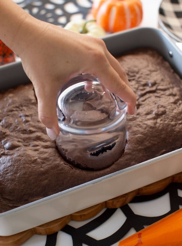 A hand presses a glass upside down into a chocolate cake in a baking pan, cutting out a circle. A small pumpkin and black-and-white tableware are visible in the background.