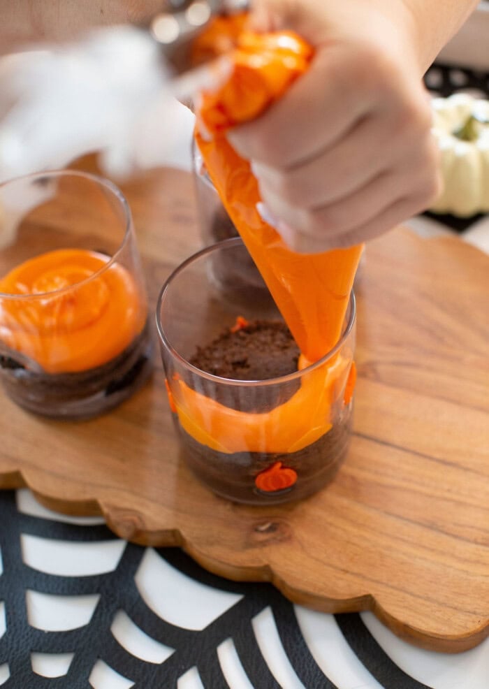 A hand pipes bright orange frosting onto a layer of chocolate cake in a glass, with more filled glasses on a wooden board and a spiderweb-patterned placemat underneath.