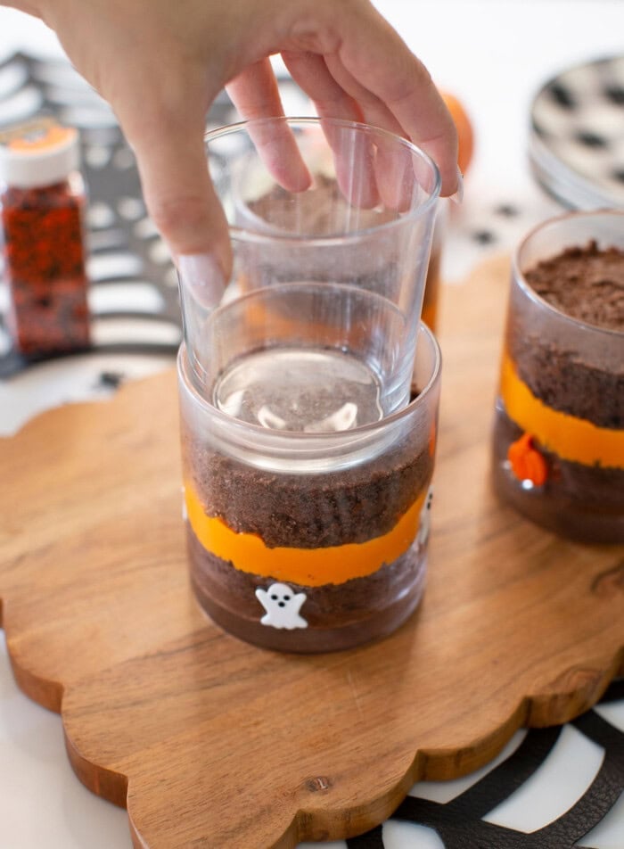 A hand presses a clear glass into a dessert layered in another glass, decorated with orange tape and ghost stickers, on a wooden board.