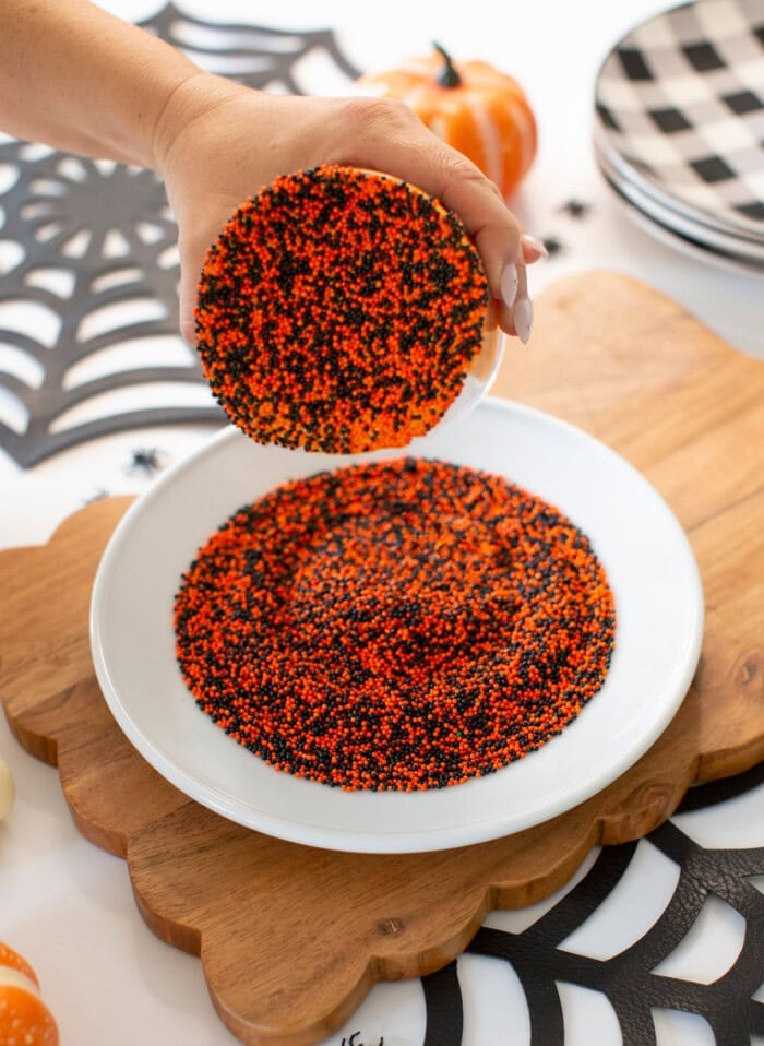 A hand dips a round cookie with orange, black, and red sprinkles onto a plate covered in matching sprinkles. Halloween-themed decor and plates are in the background.