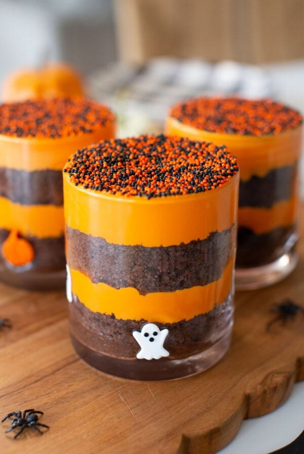 Three layered Halloween desserts in glass cups with orange and brown layers, topped with orange and black sprinkles, and decorated with ghost and pumpkin figures on a wooden board. Small plastic spiders surround the glasses.