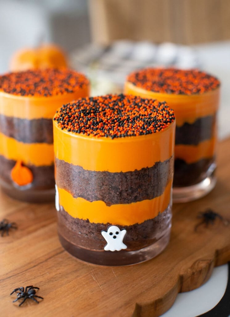 Three layered Halloween desserts in glass cups with orange and brown layers, topped with orange and black sprinkles, and decorated with ghost and pumpkin figures on a wooden board. Small plastic spiders surround the glasses.