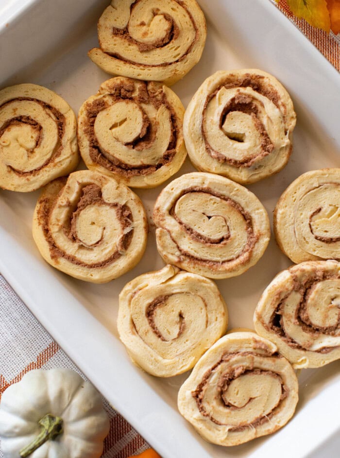 Unbaked cinnamon roll dough spirals arranged in a white rectangular baking dish, ready for baking. A small white pumpkin is visible in the bottom left corner of the image.