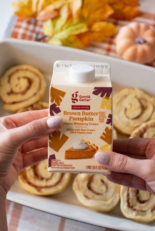 A person holds a carton of Good & Gather Brown Butter Pumpkin heavy whipping cream above a tray of unbaked cinnamon rolls, with autumn leaves and a pumpkin in the background.