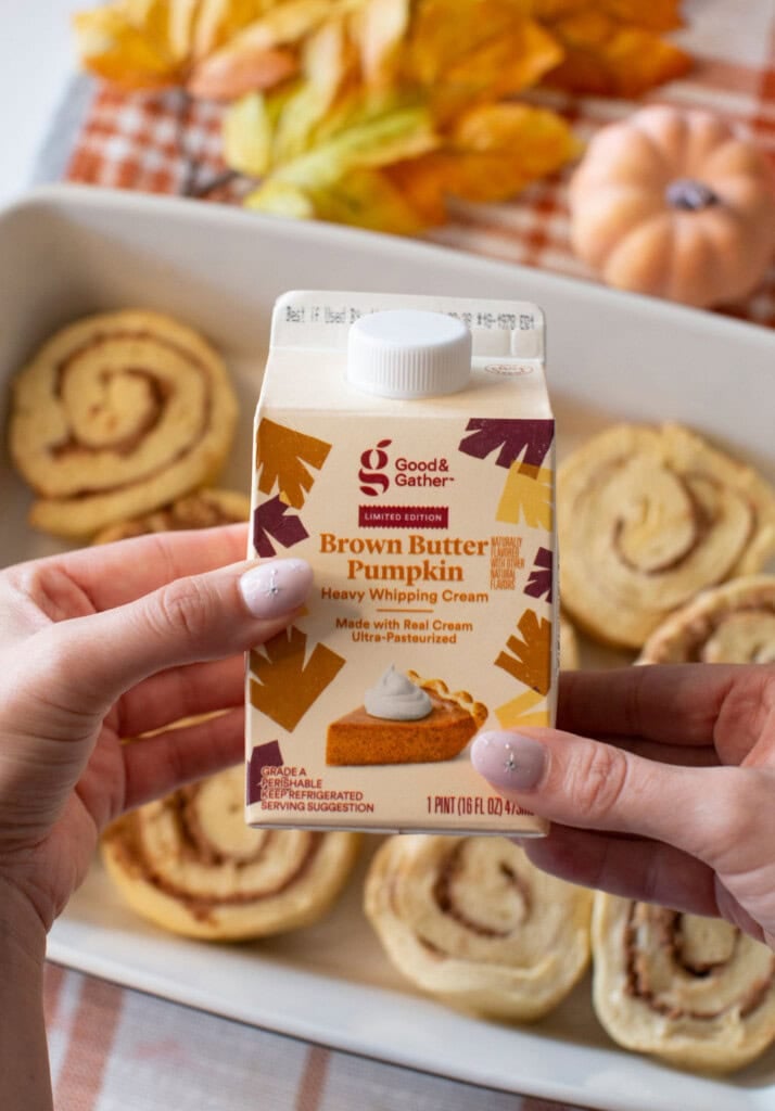 A person holds a carton of Good & Gather Brown Butter Pumpkin heavy whipping cream above a tray of unbaked cinnamon rolls, with autumn leaves and a pumpkin in the background.