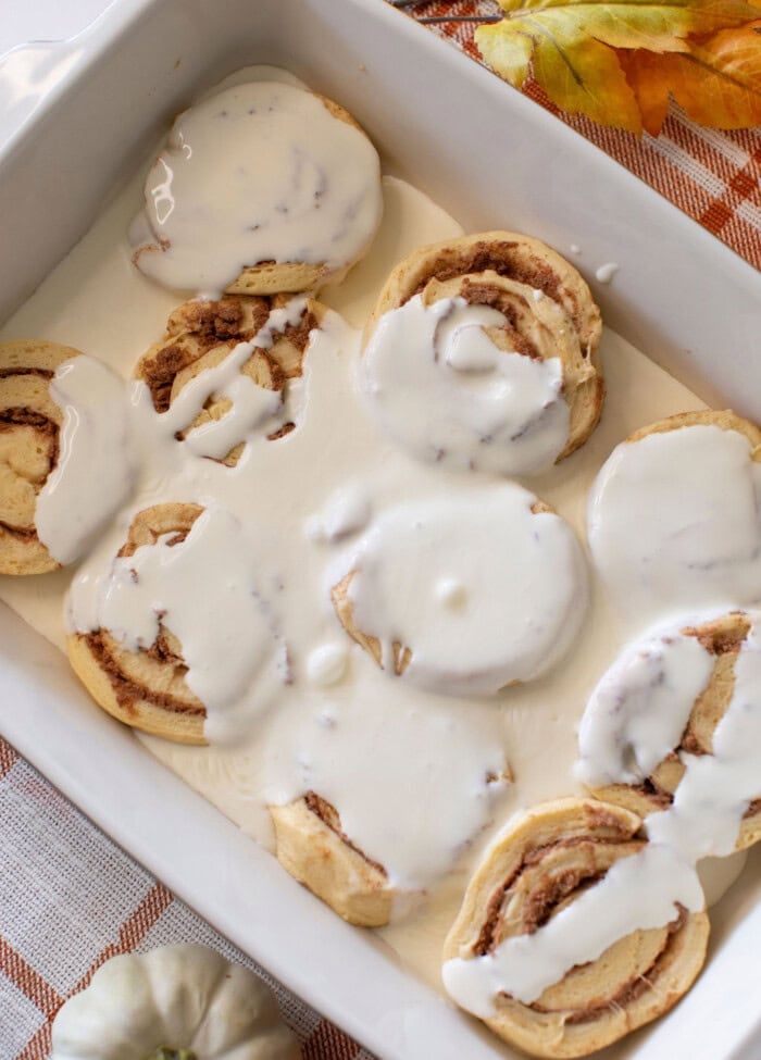 Cinnamon rolls in a white baking dish, partially covered with white icing. The dish is on a checkered orange cloth with a small white pumpkin and autumn leaves nearby.
