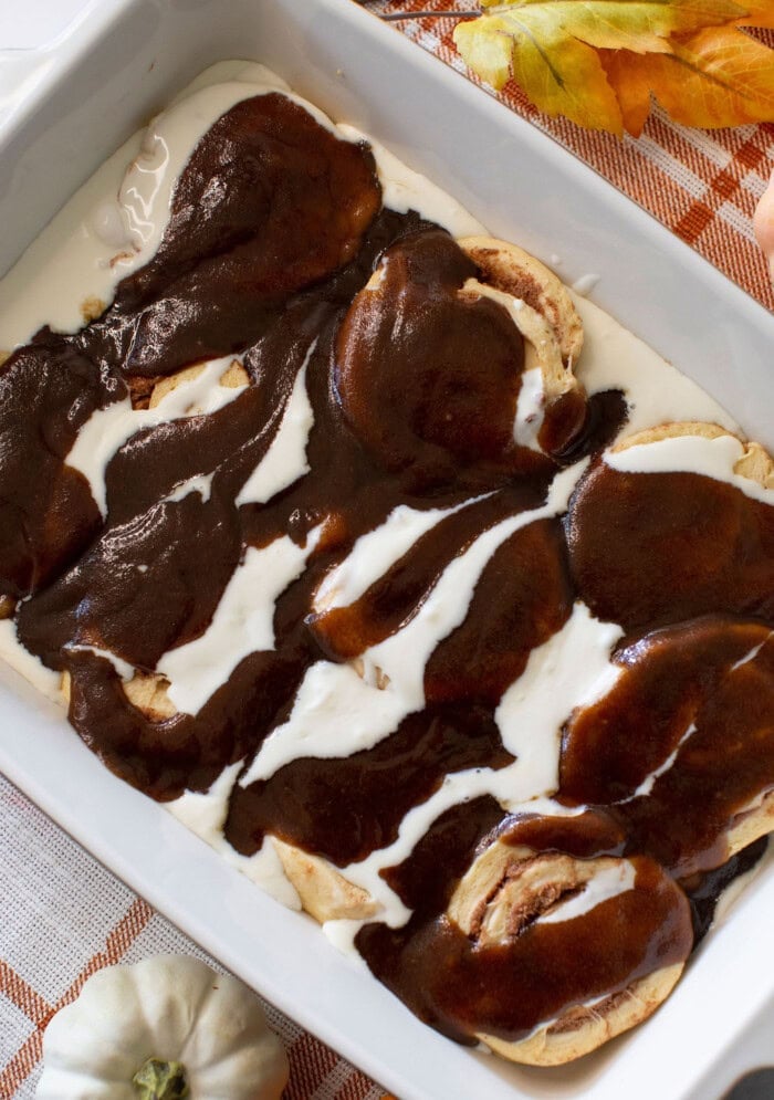 A white casserole dish filled with cinnamon rolls topped with white icing and a thick layer of dark brown chocolate sauce, set on an autumn-themed tablecloth with leaves and a small white pumpkin nearby.