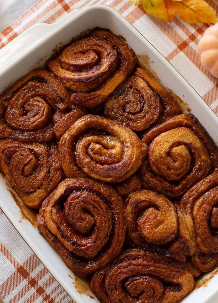 A baking dish filled with golden-brown cinnamon rolls, freshly baked and arranged closely together, sits on a plaid cloth with fall leaves nearby.