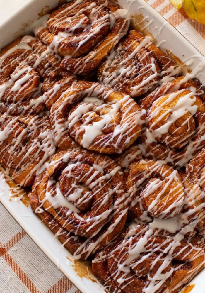 A close-up of freshly baked cinnamon rolls in a white dish, topped with a generous drizzle of white icing. The rolls are golden brown and arranged closely together.