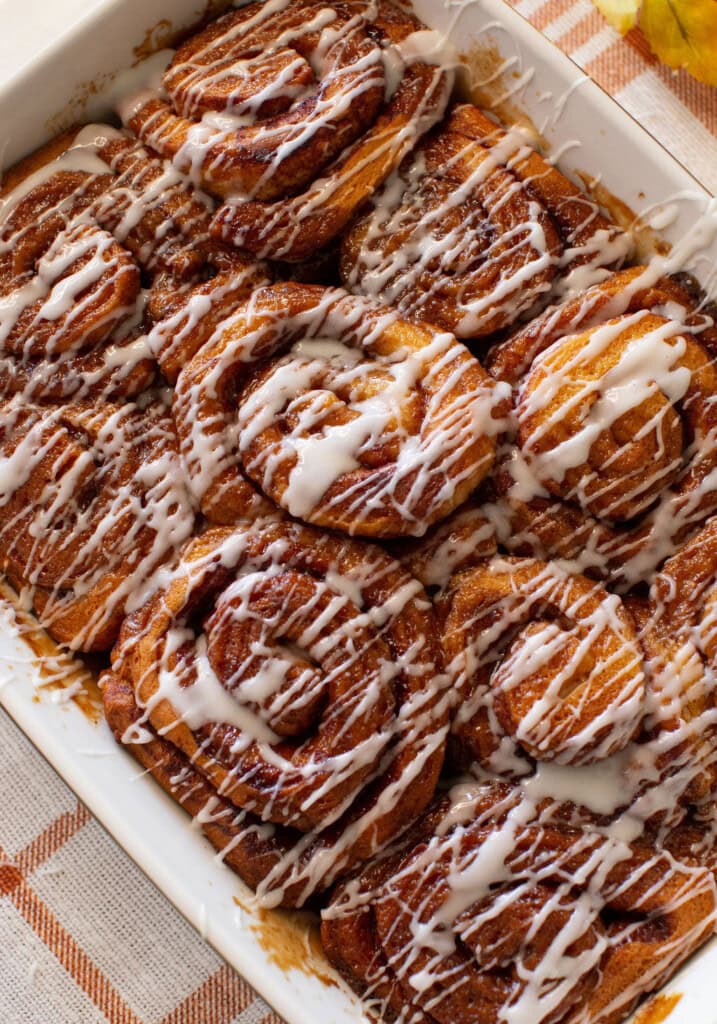 A close-up of freshly baked cinnamon rolls in a white dish, topped with a generous drizzle of white icing. The rolls are golden brown and arranged closely together.