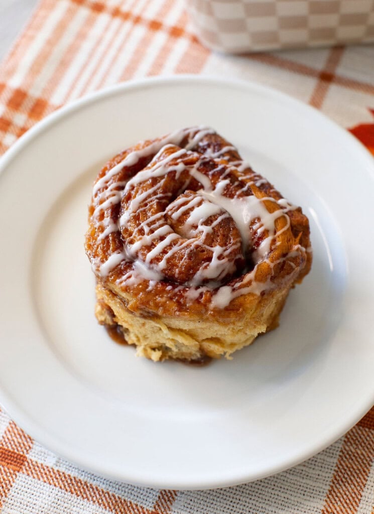 A cinnamon roll with white icing drizzle sits on a white plate atop an orange and white checkered cloth.