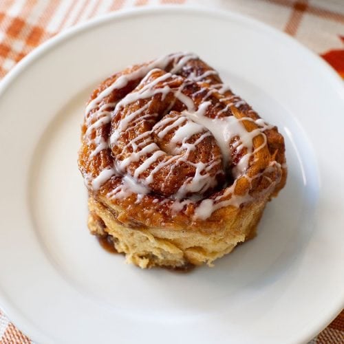 A cinnamon roll with white icing drizzled on top sits on a white plate, placed on a plaid-patterned tablecloth.