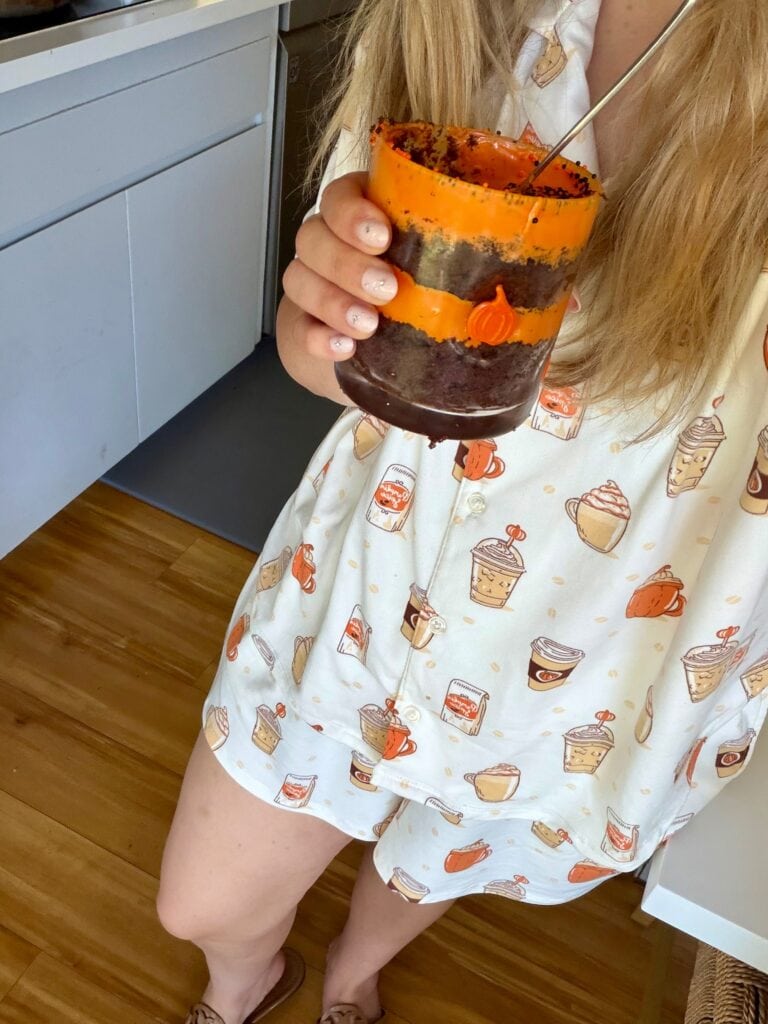 A person wearing pajamas with coffee cups holds a layered dessert in an orange cup with a spoon. Only their torso and legs are visible.