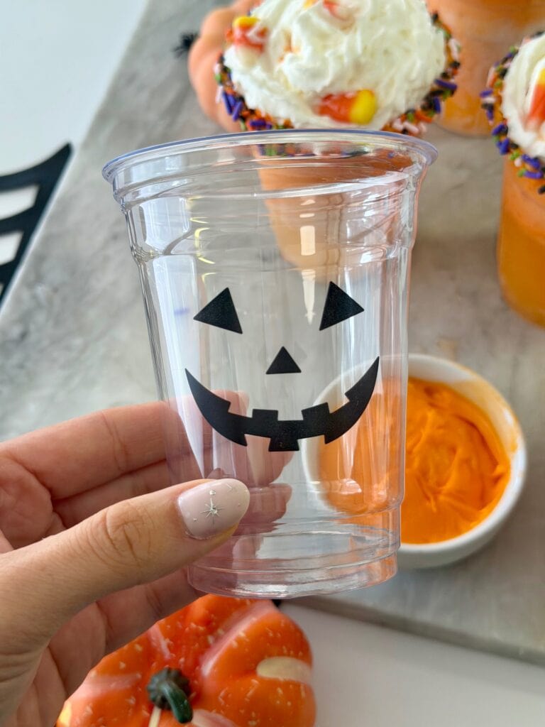 A hand holds a clear plastic cup with a black Jack-o-lantern face. In the background are orange frosted treats, a small bowl of orange frosting, and a small pumpkin.