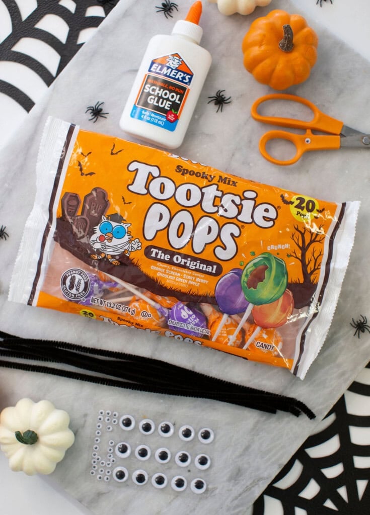 A bag of Tootsie Pops, Elmers glue, orange scissors, black pipe cleaners, googly eyes, and small white pumpkins are arranged on a white surface with black spiderweb decorations.