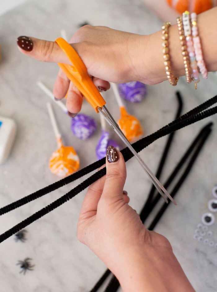 Hands using orange scissors to cut a black pipe cleaner. Colorful lollipops, small plastic spiders, and googly eyes are visible on the table below, suggesting a Halloween-themed craft activity.