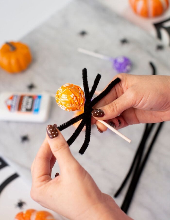 A person makes a Halloween spider craft by wrapping black pipe cleaners around an orange lollipop, with small pumpkins, glue, and another lollipop on a table in the background.
