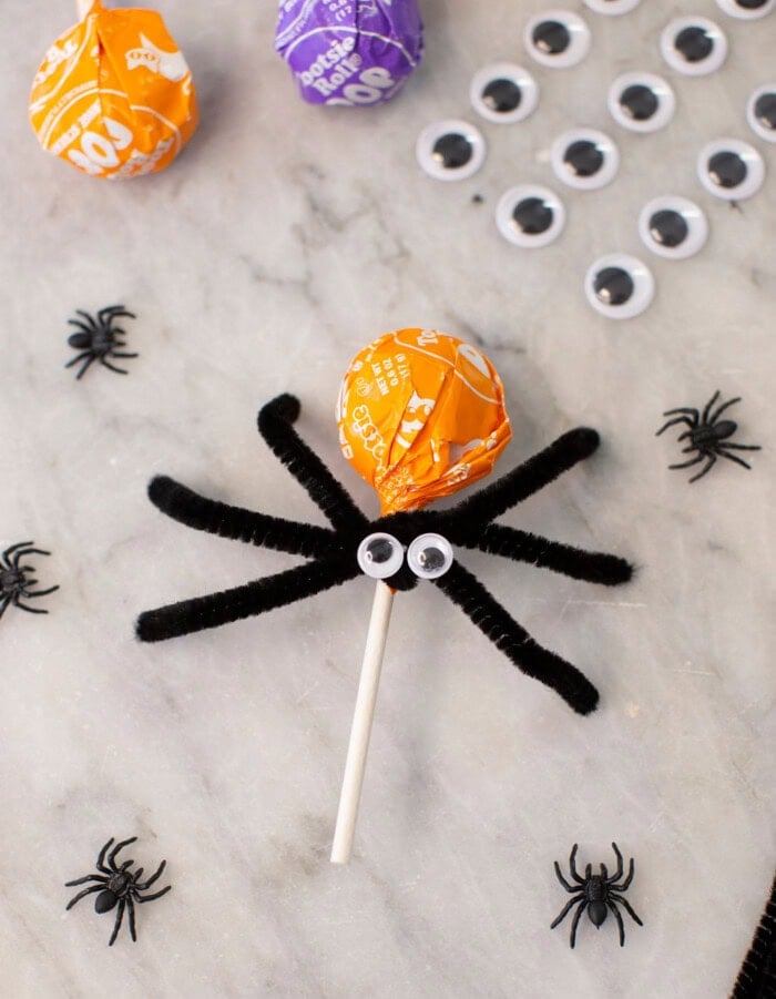 A lollipop with an orange wrapper is decorated with googly eyes and black pipe cleaners as spider legs, surrounded by plastic spiders and googly eyes on a marble surface.