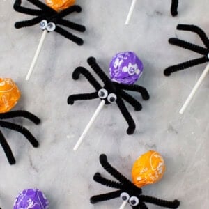 Colorful lollipops decorated as spiders with black pipe cleaner legs and googly eyes are arranged on a marble surface; a small plastic spider is also visible.