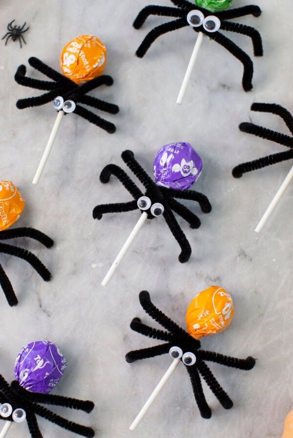 Colorful lollipops decorated as spiders with black pipe cleaner legs and googly eyes are arranged on a marble surface; a small plastic spider is also visible.