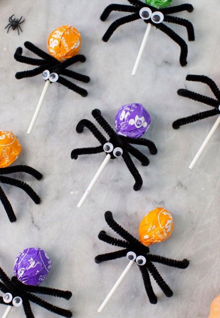 Colorful lollipops decorated as spiders with black pipe cleaner legs and googly eyes are arranged on a marble surface; a small plastic spider is also visible.